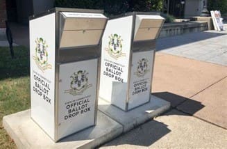 Two white ballot drop boxes sitting on the side of a road.