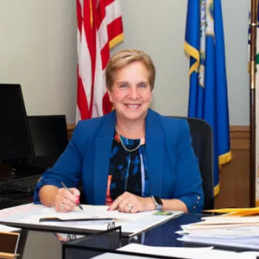 A woman sitting at a desk in front of two flags.